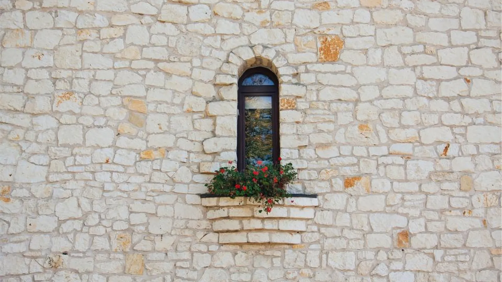 Natursteinmauer aus hellem Bruchstein mit schmalem Bogenfenster und Blumenkasten