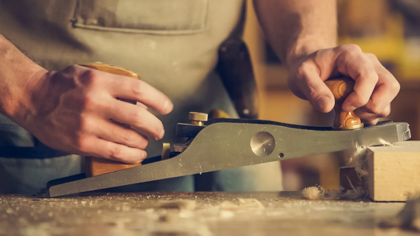 Hände führen einen Handhobel über ein Holzstück in der Werkstatt