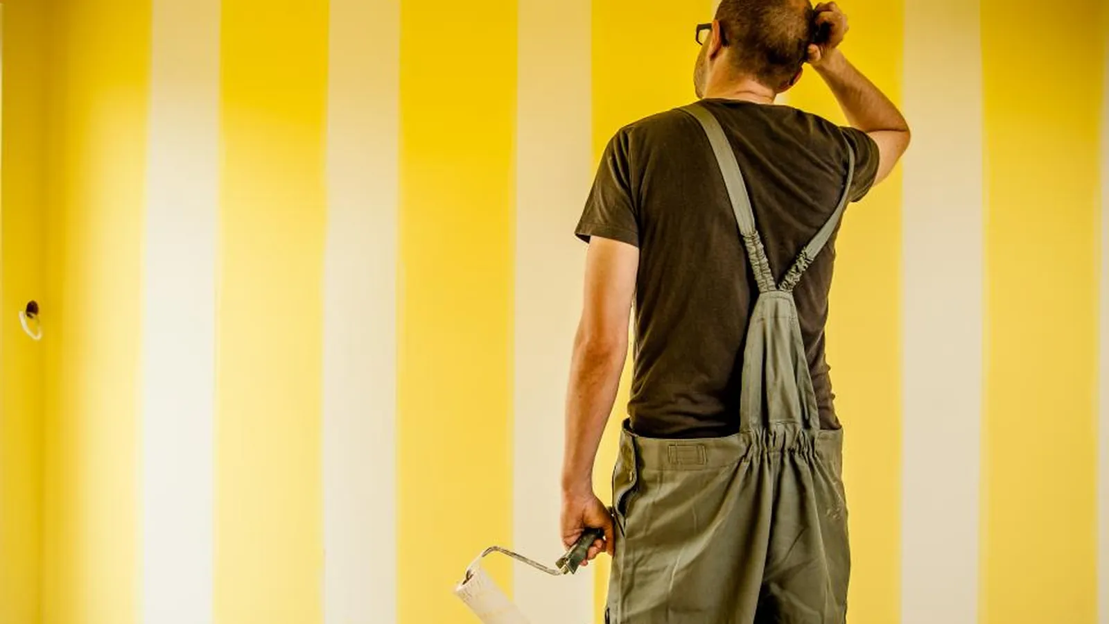 Mann in Latzhose streicht eine gelb gestreifte Wand mit der Farbrolle